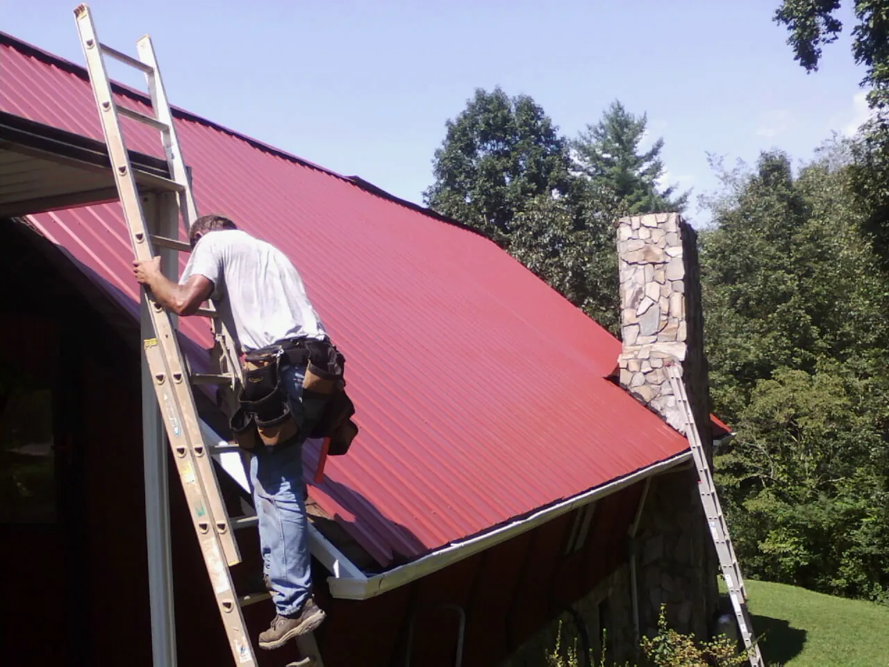 Roofer on a ladder beside a red metal roof and stone chimney — Soffit & Fascia Repair by Roofing Brothers in North Jackson